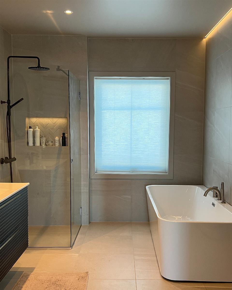 Baderom med håndbetjent topphengt duette plissegardin, hvitt badekar og dusj med glassdører. Foto.